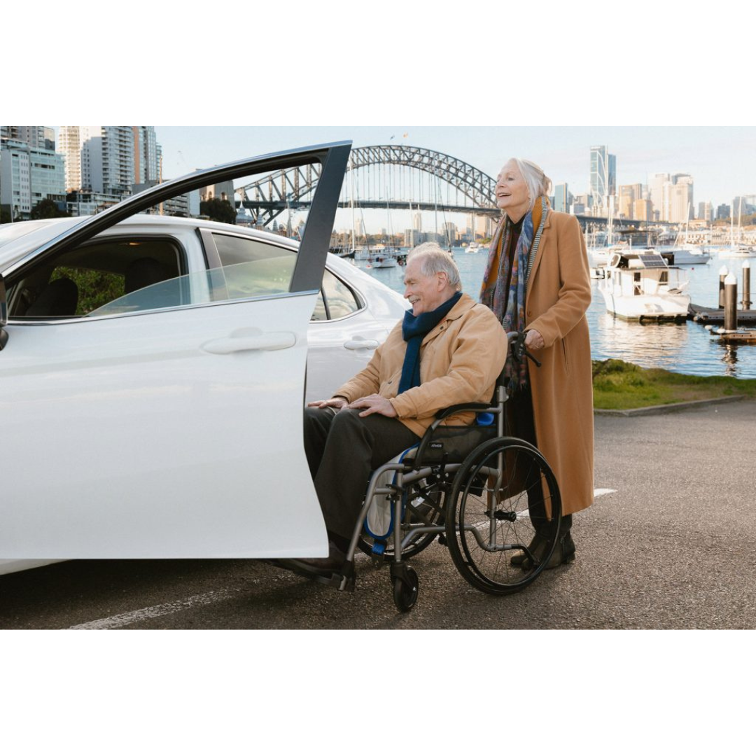 An older man in a wheelchair prepares to enter a white car using the Para Mobility IBIS Wheelchair to Car Access Lift, assisted by an older woman. They are outdoors near water with boats and a large bridge in the background, dressed in coats and scarves.