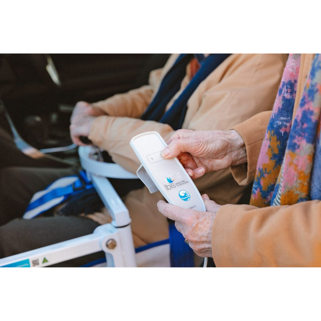 An elderly person in a tan coat holds a white electronic device labeled Idis, sitting beside someone in a wheelchair—possibly discussing the Para Mobility IBIS Wheelchair to Car Access Lift.