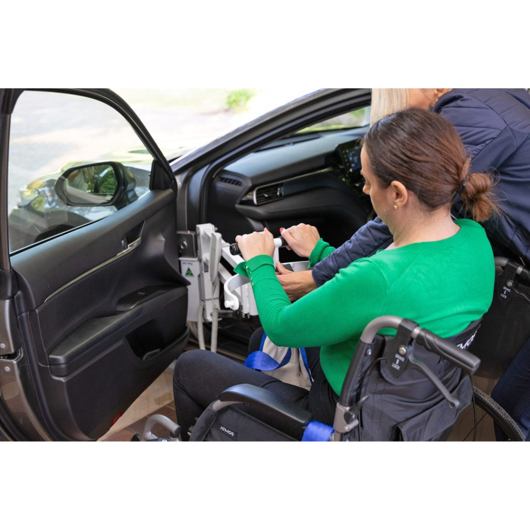 A woman in a green top sits in a wheelchair beside an open car door, assisted by another person as she prepares to use the Para Mobility IBIS Wheelchair to Car Access Lift to enter the vehicle.