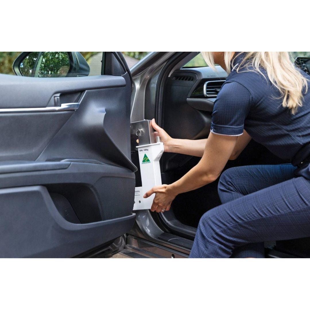 A blonde woman is installing or removing a Para Mobility IBIS Wheelchair to Car Access Lift, marked with a green warning label, from the passenger footwell of a car with its door open.