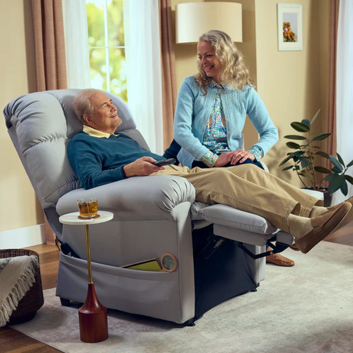 An elderly man reclines in a Journey Perfect Sleep Recliner & Lift Chair with Heat, holding a remote, as a smiling woman sits on the armrest. They relax in a cozy living room with soft lighting and massage vibrations, next to a side table with a glass of beverage.