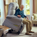 In the living room with large windows, pink curtains, and neutral furniture, an elderly man sits in a Journey Perfect Sleep Recliner & Lift Chair with Heat. The powered mechanism gently lifts him as he holds a remote control. Therapeutic heat and massage enhance his comfort.