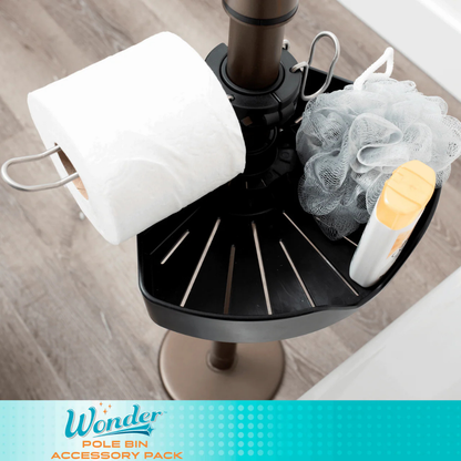 A black Stander Security Pole Bin Accessory Pack holds a toilet paper roll, a white bath pouf, and a yellow-and-white bottle, sitting on a wood floor. The bottom displays the Stander Security Pole Bin Accessory Pack branding.