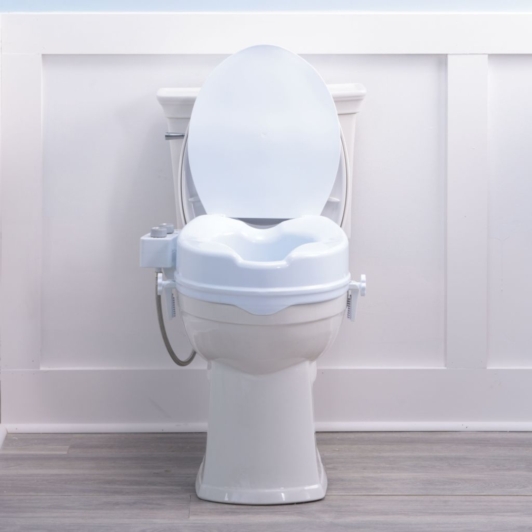 A Drive PreserveTech™ Raised Toilet Seat with Bidet - 4" Rise is shown in white, featuring an open lid. It is set against a white paneled wall and light wood flooring.