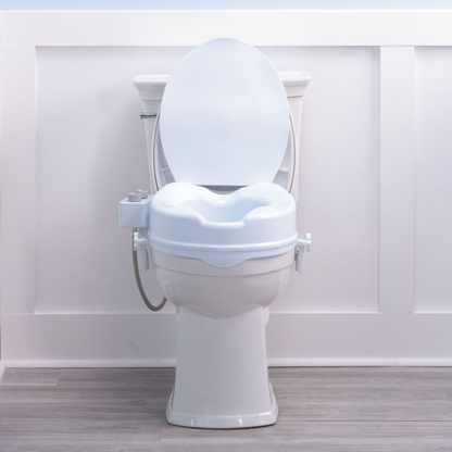 A Drive PreserveTech™ Raised Toilet Seat with Bidet - 4" Rise is shown in white, featuring an open lid. It is set against a white paneled wall and light wood flooring.