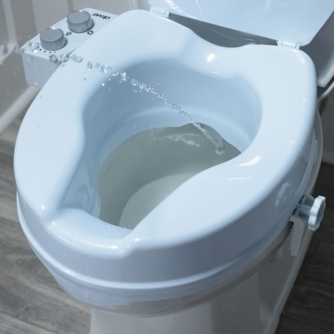 A close-up of the Drive PreserveTech™ Raised Toilet Seat with Bidet - 4" Rise shows water spraying into the bowl. The bidet’s control knobs are on the left, and the clean seat sits on a wooden floor, highlighting its modern convenience.