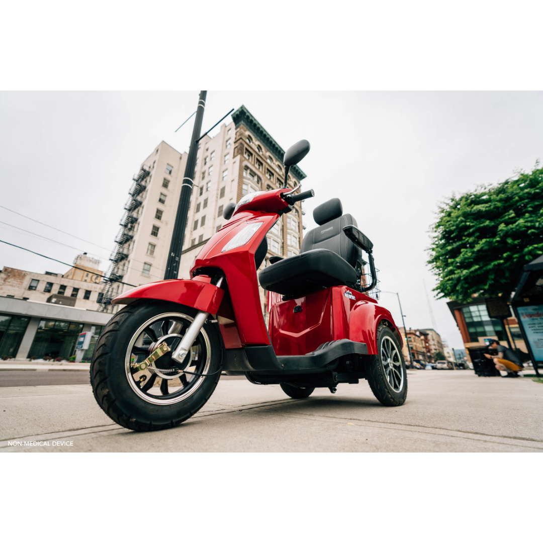 Low-angle view of a red Pride Baja® Raptor® 2 Bariatric 3-Wheel Scooter parked on a city street, featuring LED lighting, with tall buildings and trees in the background under a cloudy sky. Ideal for outdoor mobility.