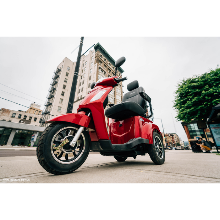Low-angle view of a red Pride Baja® Raptor® 2 Bariatric 3-Wheel Scooter parked on a city street, featuring LED lighting, with tall buildings and trees in the background under a cloudy sky. Ideal for outdoor mobility.