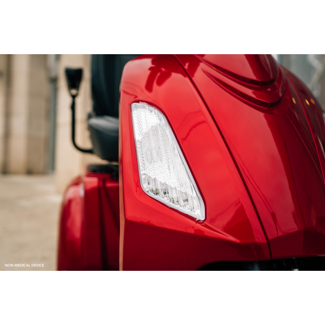 Close-up of a shiny red Pride Baja® Raptor® 2 Bariatric 3-Wheel Scooter, focusing on the front left headlight and handlebar. The blurred background highlights its sleek design and advanced full suspension for smooth outdoor rides.