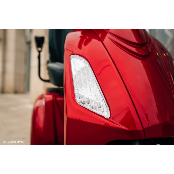 Close-up of a shiny red Pride Baja® Raptor® 2 Bariatric 3-Wheel Scooter, focusing on the front left headlight and handlebar. The blurred background highlights its sleek design and advanced full suspension for smooth outdoor rides.