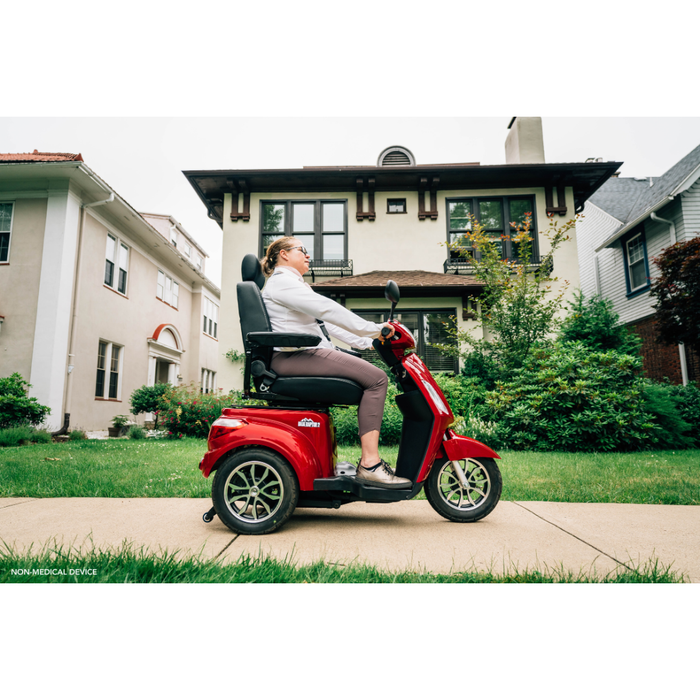A person rides a Pride Baja® Raptor® 2 Bariatric 3-Wheel Scooter with LED lighting on a sidewalk in front of a house, lawns and neighboring homes visible in the background on a cloudy day, highlighting outdoor mobility.