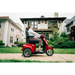 A person rides a Pride Baja® Raptor® 2 Bariatric 3-Wheel Scooter with LED lighting on a sidewalk in front of a house, lawns and neighboring homes visible in the background on a cloudy day, highlighting outdoor mobility.
