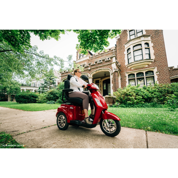 A woman rides a Pride Baja® Raptor® 2 Bariatric 3-Wheel Scooter with LED lighting on a sidewalk outside a large historic brick building, surrounded by green trees and bushes.