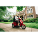 A woman rides a Pride Baja® Raptor® 2 Bariatric 3-Wheel Scooter with LED lighting on a sidewalk outside a large historic brick building, surrounded by green trees and bushes.