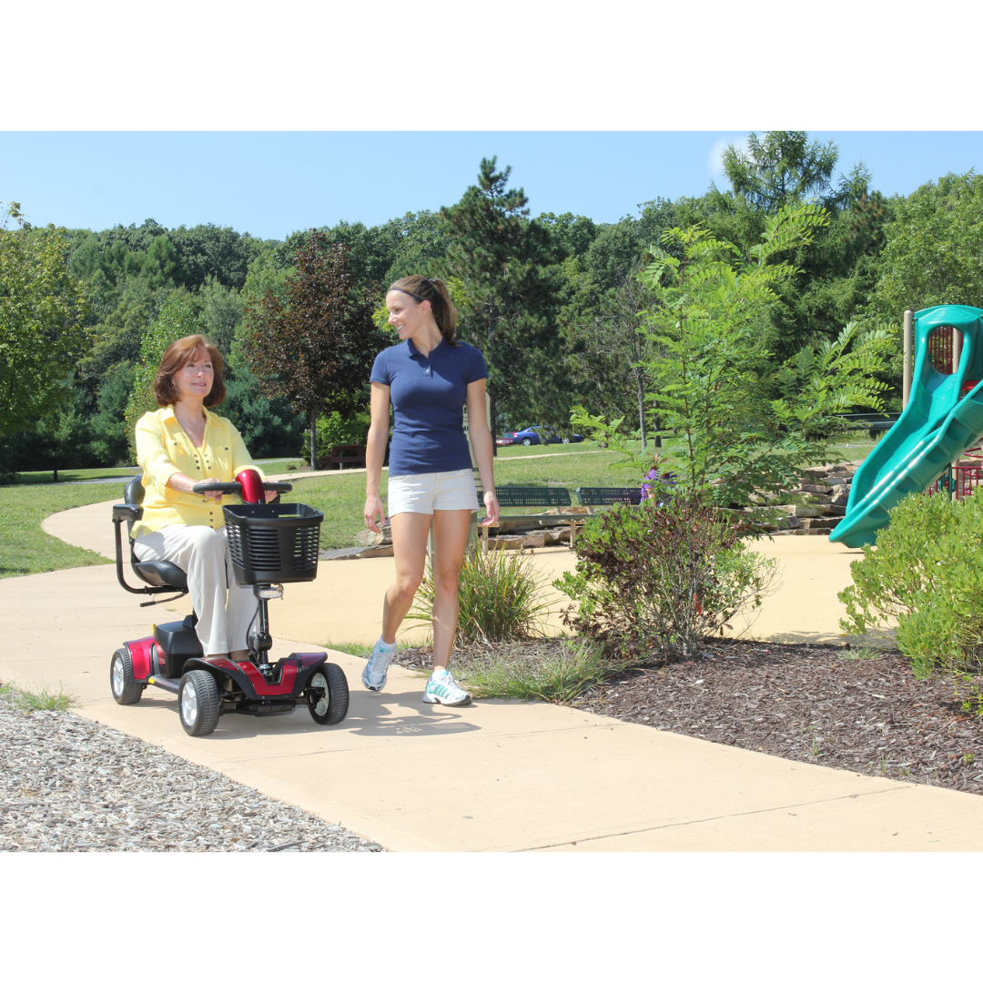 A woman in a blue shirt and shorts walks beside another on a Pride Go Go® Sport 4-Wheel Scooter along a paved path. The parks green trees, bushes, and playground slide beneath the clear sky reflect the scooters easy feather-touch disassembly for ultimate freedom.