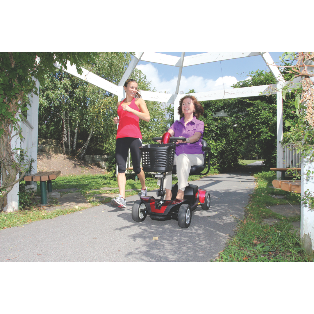 A woman in purple rides a Pride Go Go® Sport 4-Wheel Scooter along a sunny park path under a white pergola. As she goes, a woman in athletic wear jogs beside her. Trees and benches line the path, showcasing the scooter’s feather-touch disassembly for adventures like these.