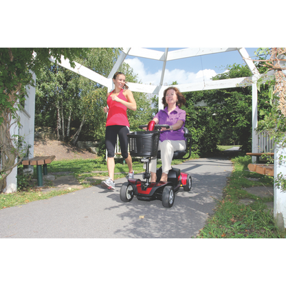 A woman in purple rides a Pride Go Go® Sport 4-Wheel Scooter along a sunny park path under a white pergola. As she goes, a woman in athletic wear jogs beside her. Trees and benches line the path, showcasing the scooter’s feather-touch disassembly for adventures like these.
