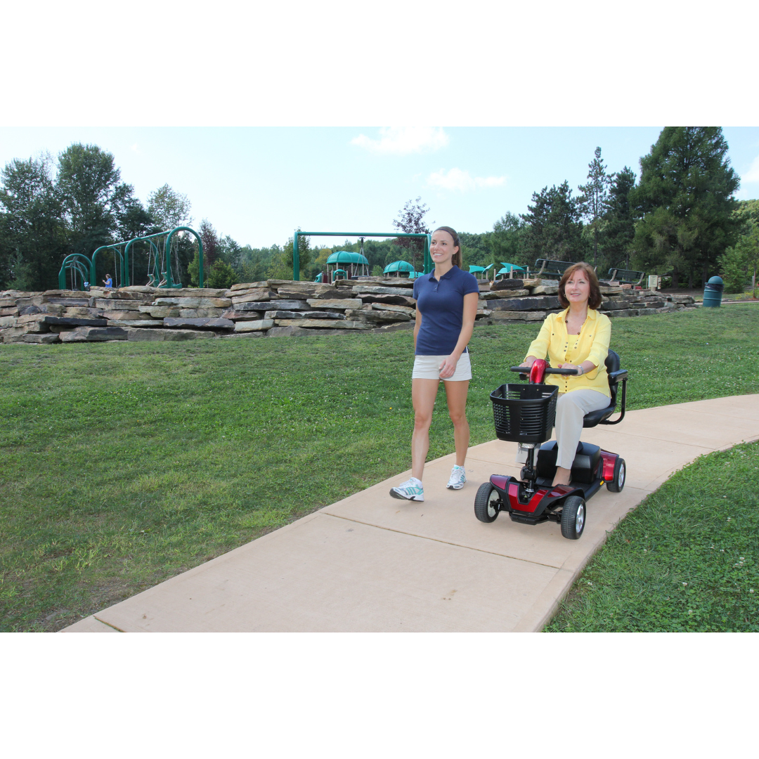 A woman rides her Pride Go Go® Sport 4-Wheel Scooter, known for its feather-touch disassembly and portability, along a paved path beside her walking companion. Enjoying the sunny park day surrounded by grass, trees, and playground equipment, both appear relaxed and content.