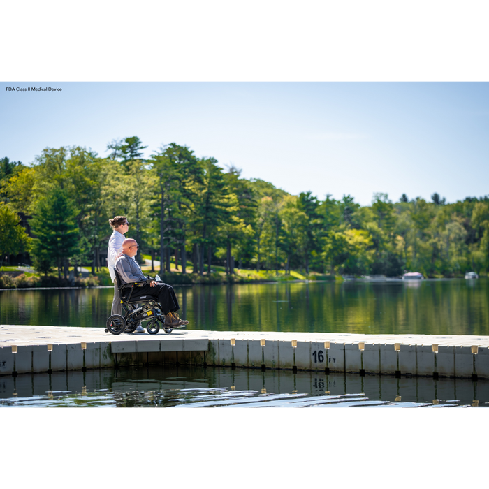 A man in a Pride Mobility Jazzy® Ultra Light Portable Electric Wheelchair sits on a lakeside dock with its carbon fiber frame shining beside another man. They both gaze over the water, surrounded by trees under the clear blue sky.
