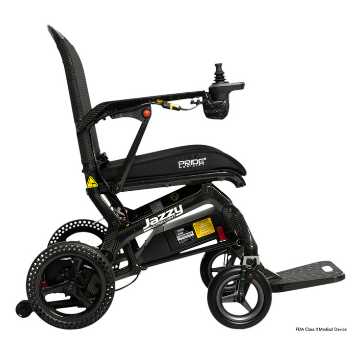 Side view of the Pride Mobility Jazzy® Ultra Light Portable Electric Wheelchair in black, featuring a carbon fiber frame, footrest, and joystick controller. It boasts a textured seat and backrest with branding and safety labels. The airline-compliant lithium battery is perfect for travel enthusiasts.