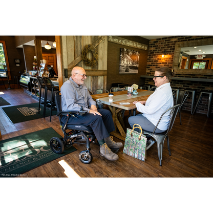 A man and woman sit at a wooden table in a cozy cafe. Beside the man is a Pride Mobility Jazzy® Ultra Light Portable Electric Wheelchair with a carbon fiber frame and airline-compliant lithium battery. A handbag rests on the floor, drinks are shared in wood and brick decor, with a counter behind them.