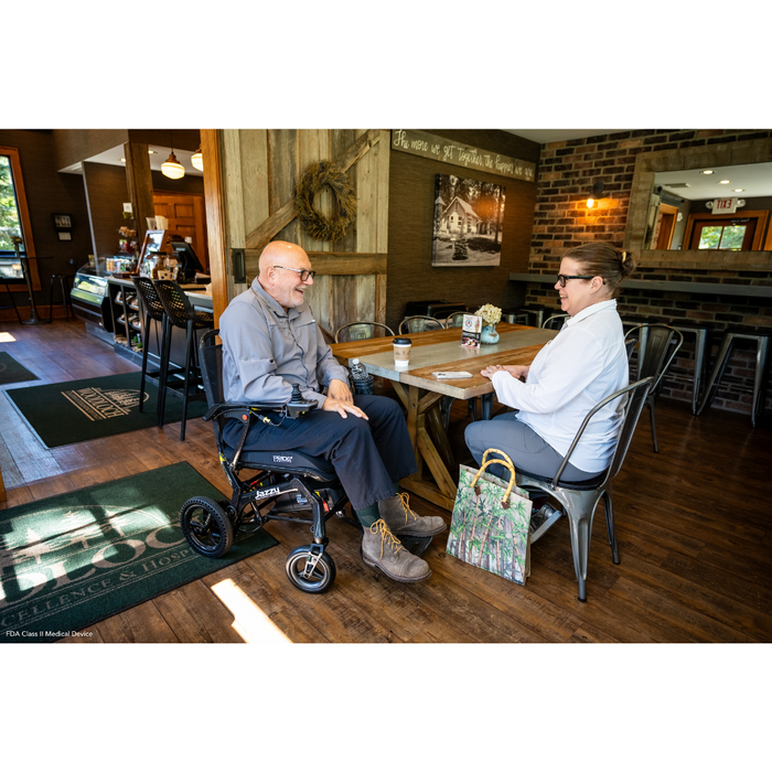 A man and woman sit at a wooden table in a cozy cafe. Beside the man is a Pride Mobility Jazzy® Ultra Light Portable Electric Wheelchair with a carbon fiber frame and airline-compliant lithium battery. A handbag rests on the floor, drinks are shared in wood and brick decor, with a counter behind them.