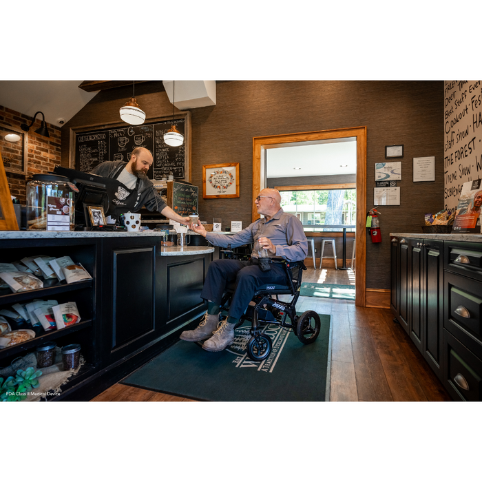 A person in a Pride Mobility Jazzy® Ultra Light Portable Electric Wheelchair receives a drink from a barista at a café counter. The cozy interior, with brick and wood walls, complements the stylish wheelchairs carbon fiber frame as it maneuvers through menu displays with ease.