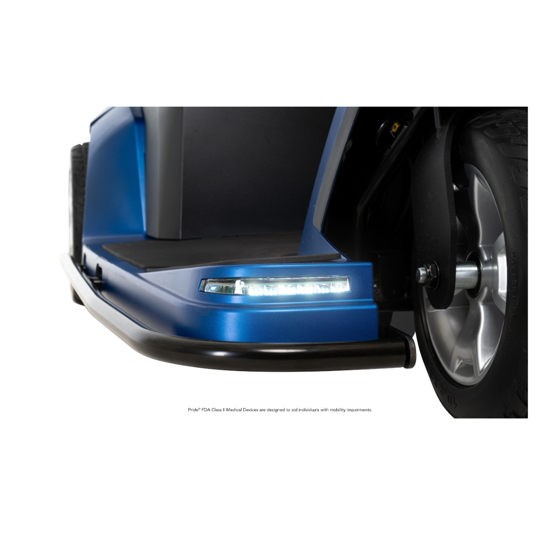 Close-up of the Pride Mobility Maxima 3-Wheel Bariatric Scooter’s front corner, highlighting the footrest platform, LED headlight, black bumper, and part of a large front wheel on a white background. Supports up to 500 lbs.