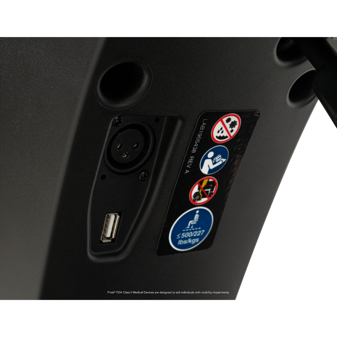 Close-up of the Pride Mobility Maxima 4-Wheel Bariatric Scooter control panel, featuring an XLR port, USB port, and safety labels showing a 500 lbs/227 kgs weight capacity plus warnings against liquids and tampering.
