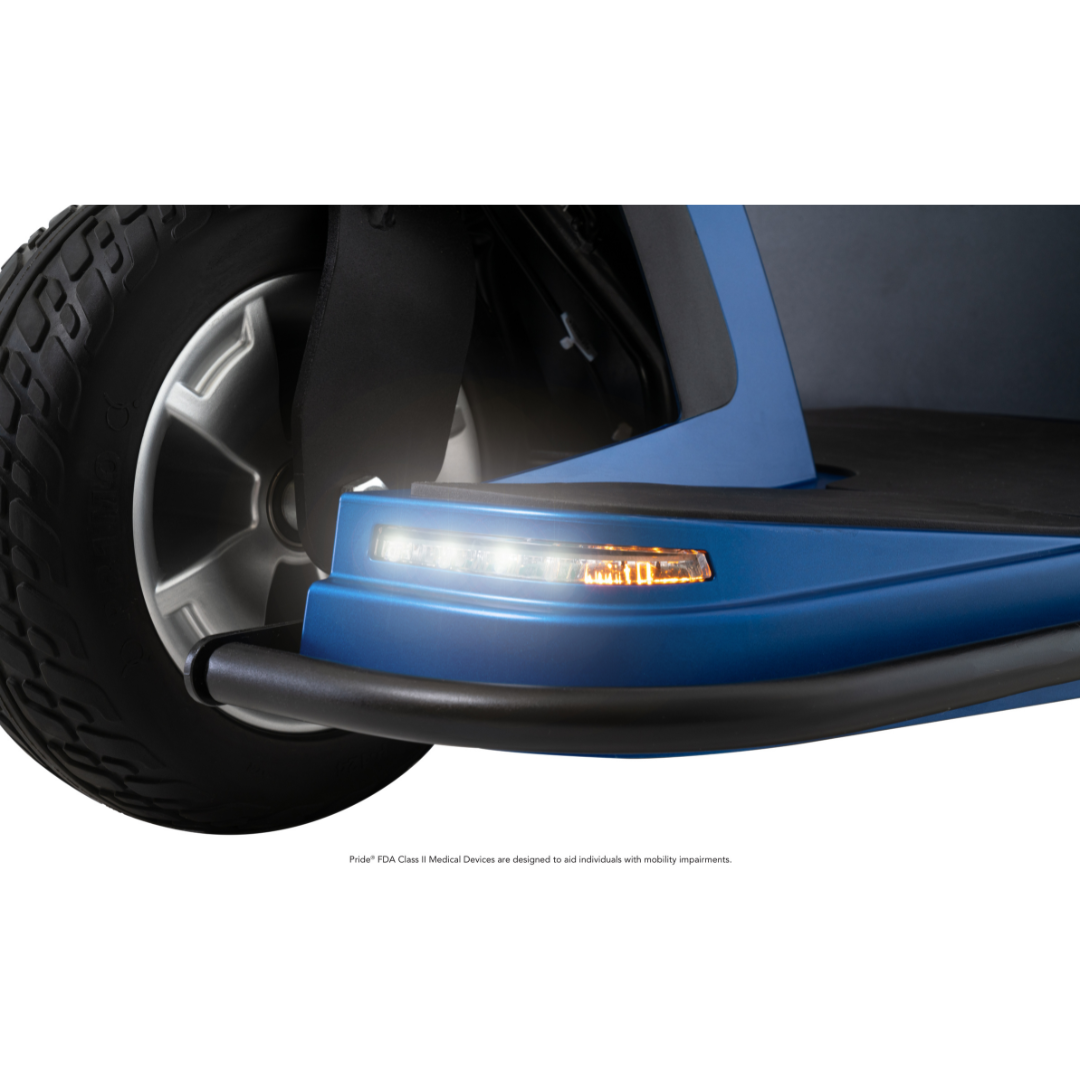 Close-up of the Pride Mobility Maxima 3-Wheel Bariatric Scooter’s front tire and blue fender, showcasing an LED headlight, orange indicator light, and black bumper edge. This scooter supports up to 500 lbs with heavy-duty performance.