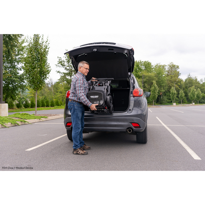 In a parking lot, a man is loading a portable lift device and the Pride Mobility GoGo Carbon Ultralite Mobility Scooter, weighing just 35 lbs, into the trunk of an SUV parked on an empty road. The vehicle is surrounded by swaying grass and towering trees beneath an overcast sky.
