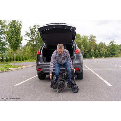 A man in a plaid shirt unloads his Pride Mobility GoGo Carbon Ultralite Scooter, weighing only 35 lbs, from the trunk of a gray SUV in an empty parking lot. With trees and clouds framing the scene, he appreciates its lightweight design for easy transport.