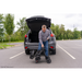 In a parking lot, a man in a plaid shirt and jeans unloads the Pride Mobility GoGo Carbon Ultralite Mobility Scooter—just 35 lbs—from the trunk of a parked grey SUV. A backdrop of green trees surrounds the scene.
