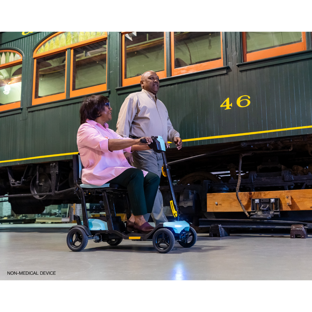 A woman rides a Pride Mobility Go Go® Super Portable Mobility Scooter as a man walks beside her in a museum, with a vintage tram adorned in green and yellow details in the background. Both are smiling.