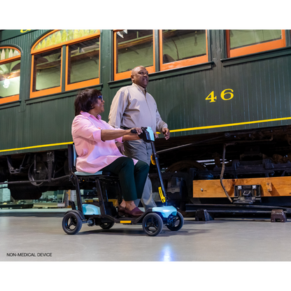 A woman rides a Pride Mobility Go Go® Super Portable Mobility Scooter as a man walks beside her in a museum, with a vintage tram adorned in green and yellow details in the background. Both are smiling.