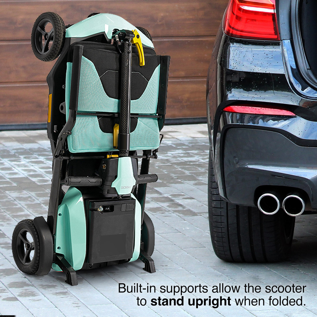 A folded Pride Mobility Go Go® Super Portable Mobility Scooter with built-in supports stands upright on a driveway next to a car. The lightweight scooter is light green and black, with a garage door and the rear of the car visible in the background.