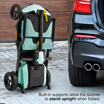 A folded Pride Mobility Go Go® Super Portable Mobility Scooter with built-in supports stands upright on a driveway next to a car. The lightweight scooter is light green and black, with a garage door and the rear of the car visible in the background.