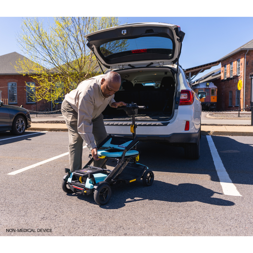 An elderly man assembles his Pride Mobility Go Go® Super Portable Mobility Scooter behind his car in a parking lot. The trunk is open, showing an airline-approved battery, as sunlight gently illuminates nearby trees and buildings.