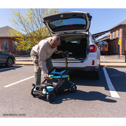 An elderly man assembles his Pride Mobility Go Go® Super Portable Mobility Scooter behind his car in a parking lot. The trunk is open, showing an airline-approved battery, as sunlight gently illuminates nearby trees and buildings.