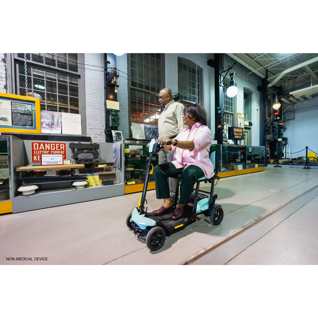 A man and a woman effortlessly glide through an industrial-themed museum on the Pride Mobility Go Go® Super Portable Mobility Scooter. Surrounded by vintage equipment and safety signs, they smoothly traverse the floor under warm lighting, powered by an airline-approved battery.