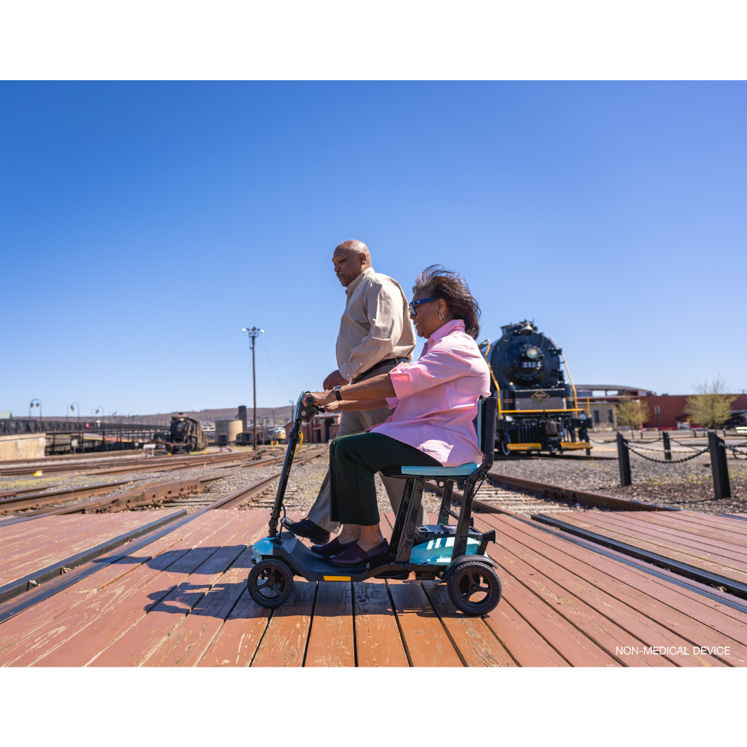 An elderly couple delights in a sunny day on a wooden platform near railway tracks. The woman sits on her Pride Mobility Go Go® Super Portable Mobility Scooter as the man strolls nearby, with a majestic steam locomotive enhancing the nostalgic ambiance of the charming scene.