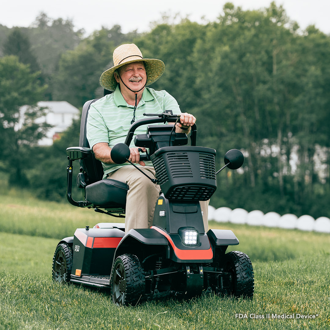 An older man in a sun hat and polo shirt smiles while driving the Pride Mobility PX4 HD Bariatric Scooter across a grassy field, with trees and a house in the background.