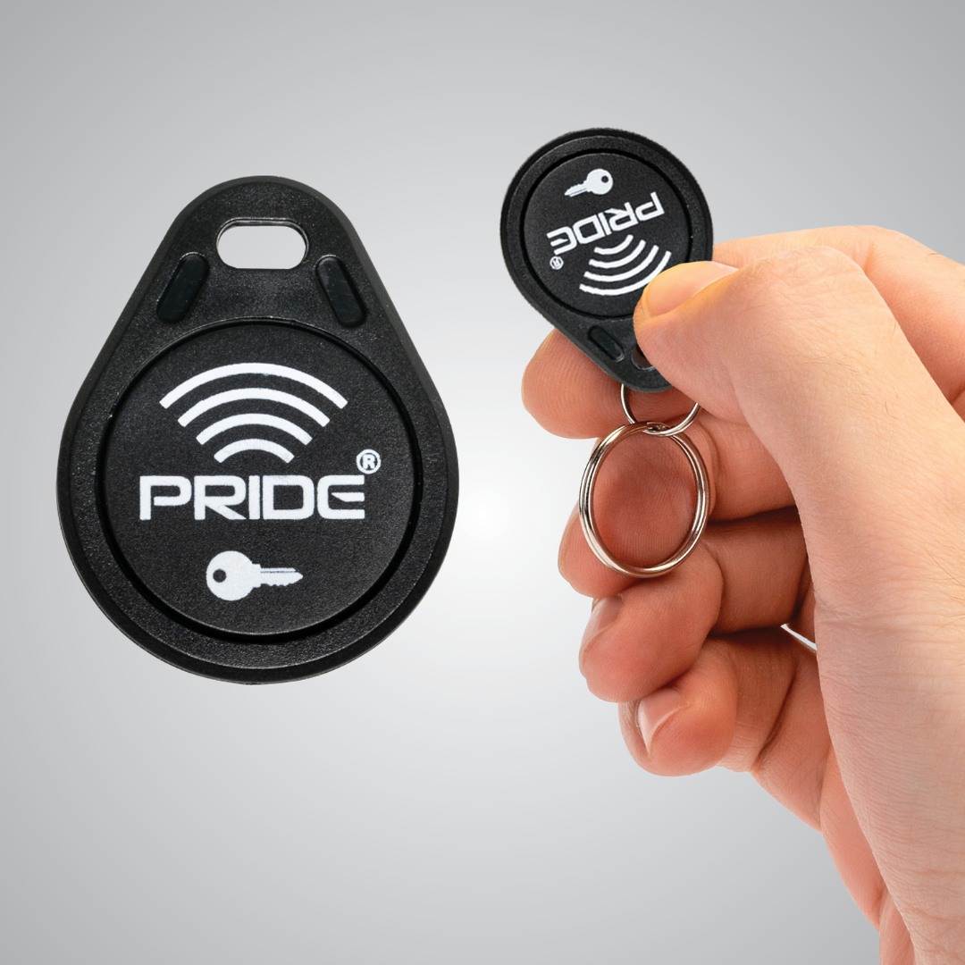 A hand holds a black RFID key fob labeled PRIDE with a key symbol, designed for the Pride Mobility PX4 HD Bariatric Scooter. Another identical key fob is shown on the left against a gray gradient background.