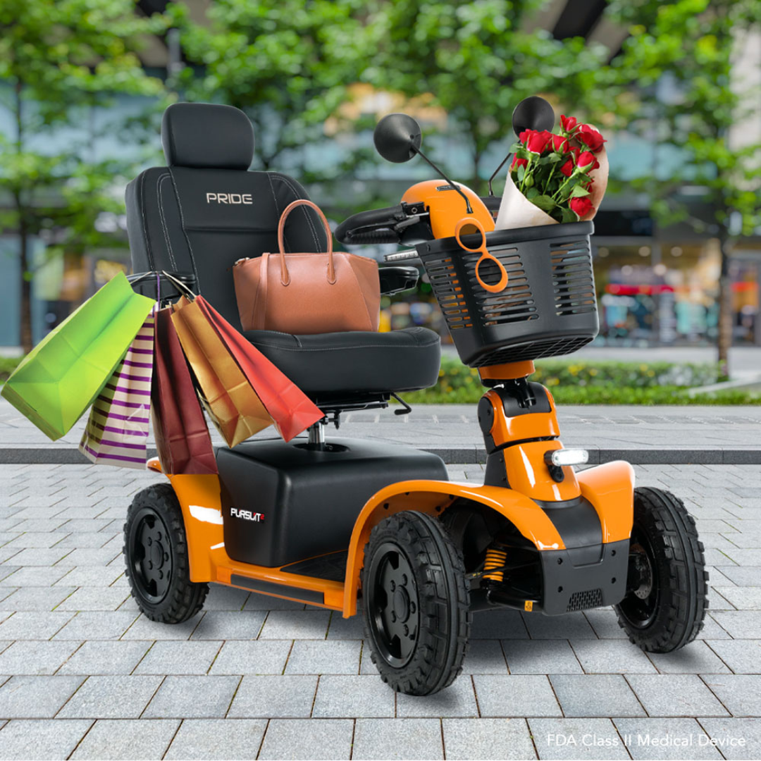 A Pride Mobility Pursuit® 2 Offroad HD Scooter in orange sits on a city sidewalk, featuring a basket with a purse and flowers, and several colorful shopping bags hanging from its armrest.