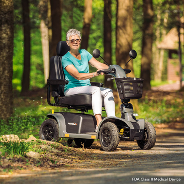 An older woman with short gray hair, glasses, and a turquoise shirt smiles while sitting on a Pride Mobility Pursuit® 2 Offroad HD Scooter along a wooded path as sunlight filters through the trees.