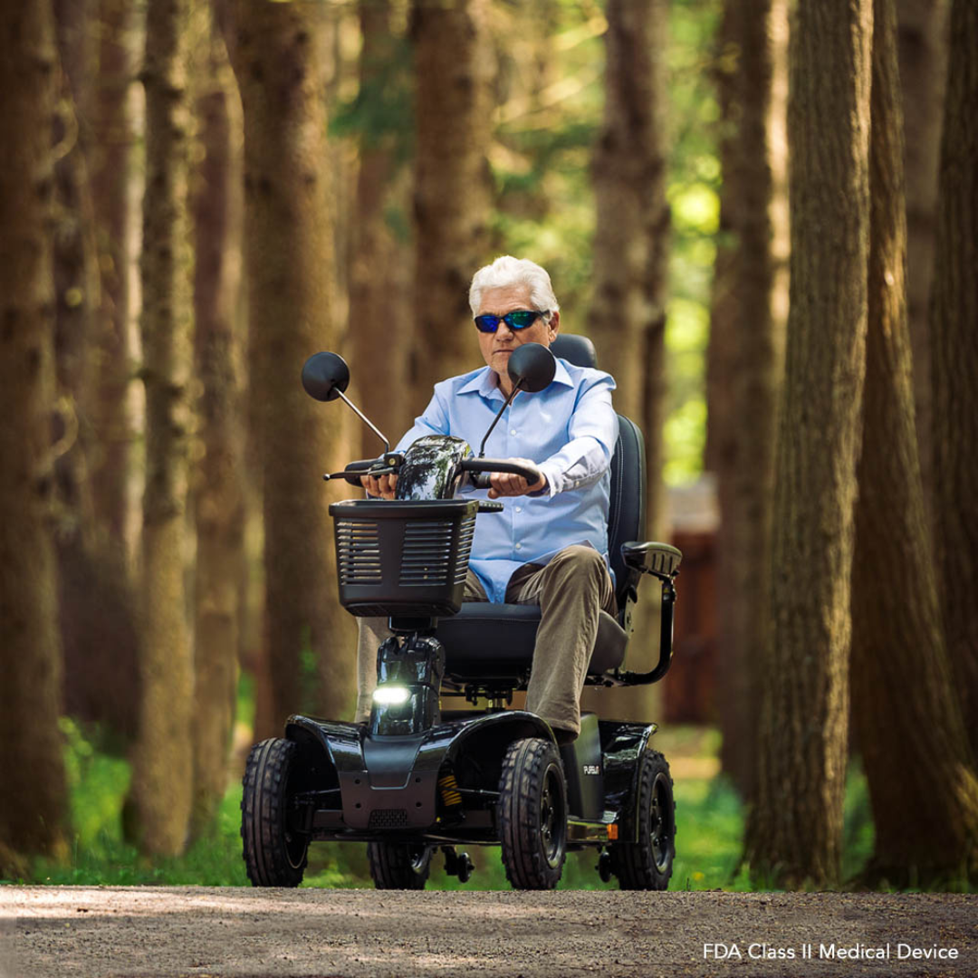 An older man wearing sunglasses rides the Pride Mobility Pursuit® 2 Offroad HD Scooter along a wooded path, surrounded by tall trees and sunlight streaming through the forest.