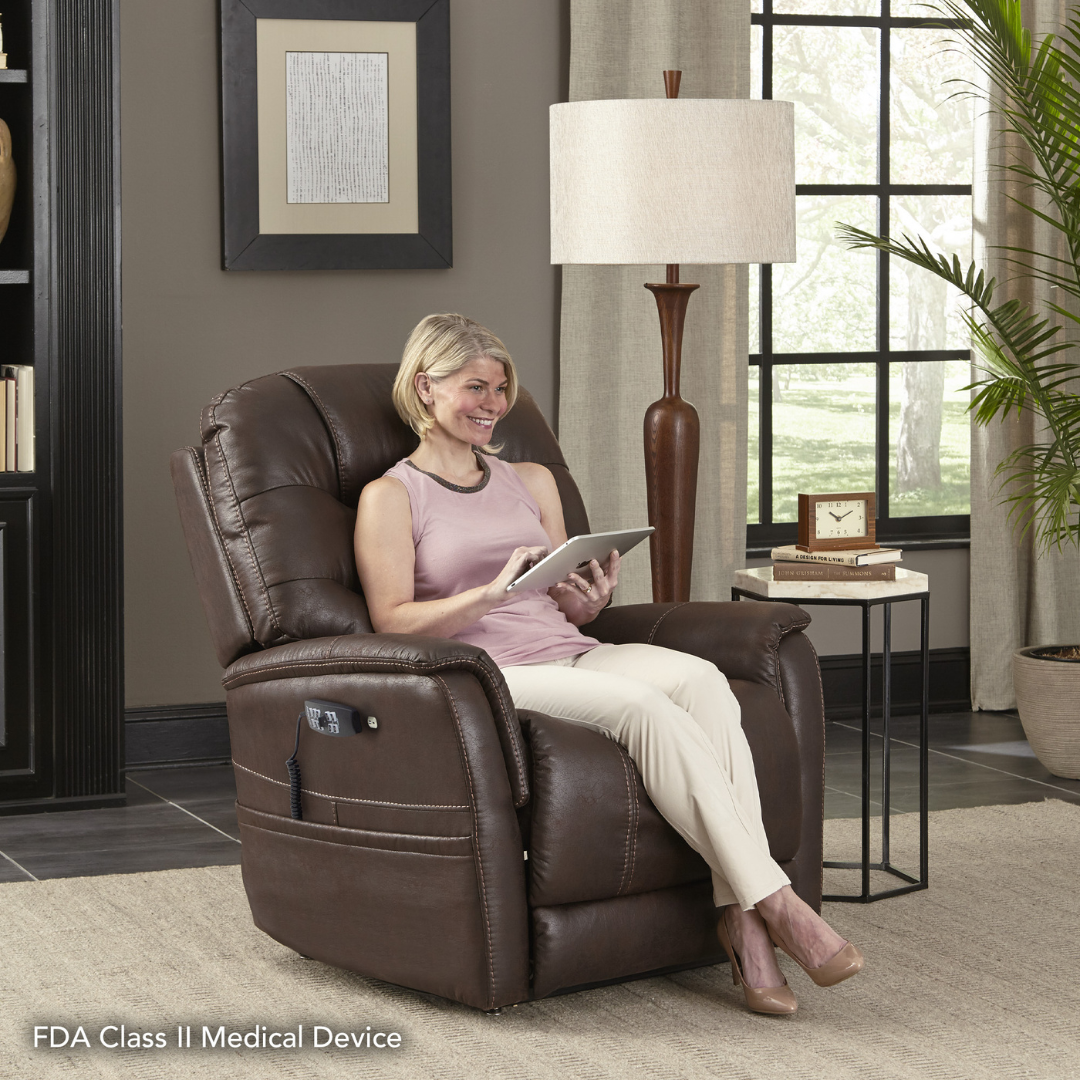 A woman relaxes in a brown Pride VivaLift!® Elegance 2 Recliner with Lift Assist, smiling with her tablet. The chair’s controls appear in a stylish living room. Text: FDA Class II Medical Device.