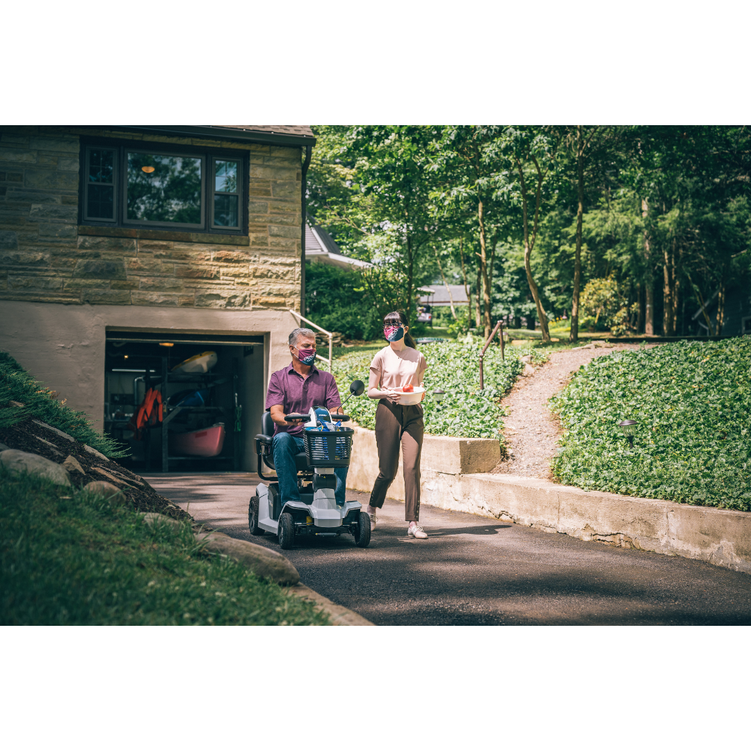 An older man rides a Pride Mobility Zero Turn 10 HD Scooter up a driveway next to a house, accompanied by a young woman walking beside him. They enjoy a conversation together on a sunny day with greenery all around.