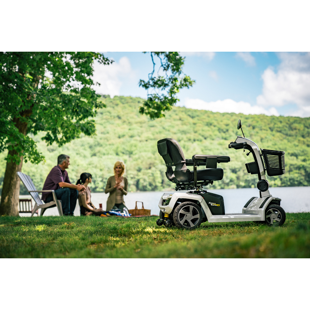 A Pride Mobility Zero Turn 10 HD Scooter is parked on grass near a lake, while three people chat at a picnic in the background. Trees and hills can be seen under a partly cloudy sky.