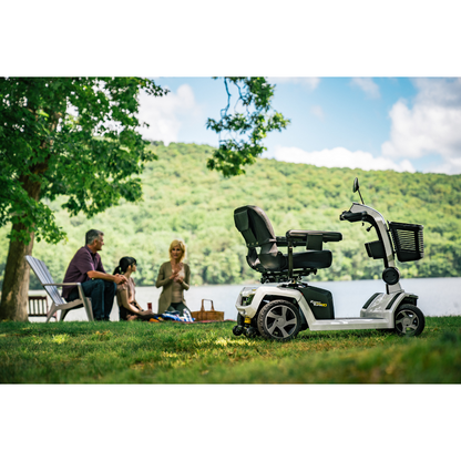 A Pride Mobility Zero Turn 10 HD Scooter is parked on grass near a lake, while three people chat at a picnic in the background. Trees and hills can be seen under a partly cloudy sky.